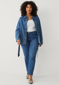 Denimjas met een relaxte pasvorm, knopen aan de voorkant en zijzakken, gedragen over een wit tanktop. Gekleed met blauwe jeans en gestreepte schoenen.