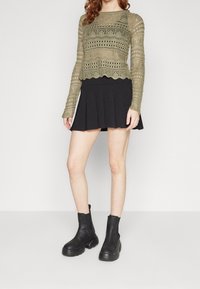 Olivenfarbene Strickjacke mit offenen Mustern und gezacktem Saum, kombiniert mit einem schwarzen, gefalteten Minirock und schwarzen Ankle Boots.