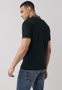 Juodas polo marškinėlis klasikinio dizaino, su tekstūruotu apykakliu ir auksiniais akcentais. Dėvimas su šviesiai pilkais džinsais.