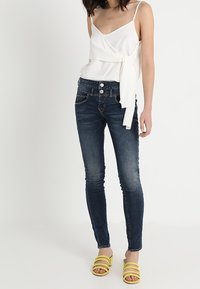 Vit omslagstopp med sidoknytning, mörkblå skinny jeans och gula snörsandaler. Släta texturer och figurnära silhuett.