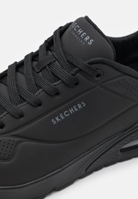 Black athletic shoe with textured upper, featuring flat laces, perforated accents, and "SKECHERS" branding on the side.