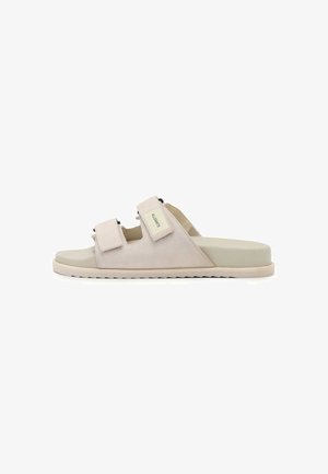 Lichtbeige slipper met twee verstelbare klittenbandsluitingen en platte zool, zijaanzicht op witte achtergrond.