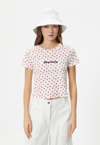 Top corto de manga corta blanca con lunares rojos, con la palabra "Fresa" en cursiva negra. Combinado con pantalones cargo blancos y un sombrero tipo cubo.