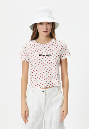 Top corto de manga corta blanca con lunares rojos, con la palabra "Fresa" en cursiva negra. Combinado con pantalones cargo blancos y un sombrero tipo cubo.
