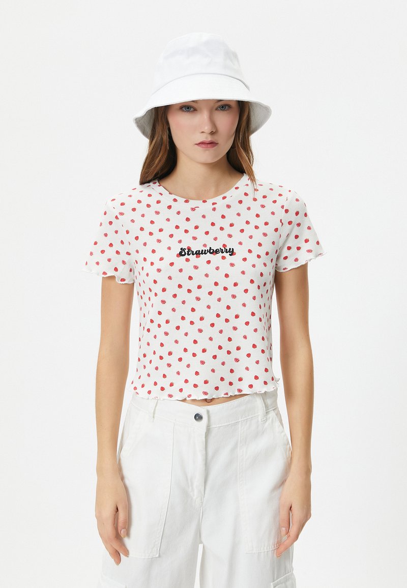 Top corto de manga corta blanca con lunares rojos, con la palabra "Fresa" en cursiva negra. Combinado con pantalones cargo blancos y un sombrero tipo cubo.