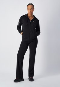 Sudadera negra con cremallera y cordón, combinada con pantalones negros a juego. La sudadera presenta un logo en el pecho y en la manga; los pantalones tienen un pequeño logo en el dobladillo.