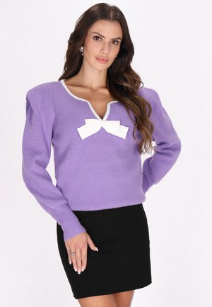 Lila gestrickter Pullover mit V-Ausschnitt und puffigen Ärmeln, versehen mit einer weißen Schleifenakzent am Brustbereich, kombiniert mit einem schwarzen Rock.