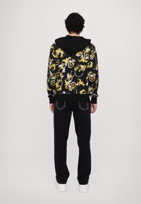 Μαύρη κουκούλα με σχέδιο κίτρινου και λευκού στροβιλισμού, διαθέτει μπροστινή τσέπη και ριμπ μανσέτες, συνδυασμένη με μαύρο παντελόνι και λευκά αθλητικά παπούτσια.