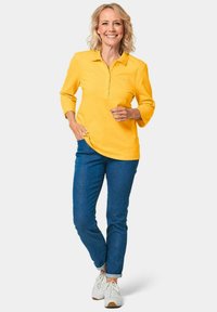 Geel katoenen poloshirt met driekwart mouwen, voorzien van een vooroverlap en een gebogen zoom, gecombineerd met blauwe jeans en witte sneakers.
