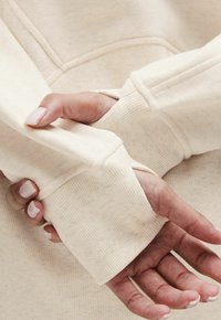 Hanorac de culoare crem, cu manșete cu nervuri. Materialul pare moale, cu o textură subtilă. Designul include un buzunar tip cangur și detalii de cusătură.