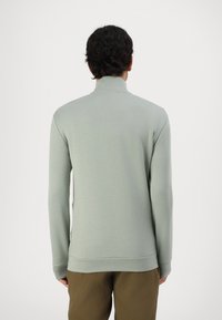 Sudadera verde claro con cuello alto, mangas largas y puños acanalados, que presenta una textura suave y una etiqueta de marca sutil en el dobladillo.