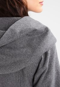 Šedá vlnená mikina s širokou, měkkou kapucí. Má hladkou texturu a skládací límcový detail, který zdůrazňuje řemeslné zpracování.