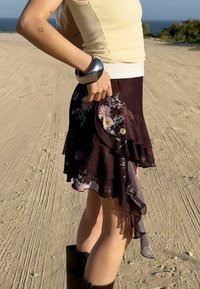 Jupe noire florale à couches avec volants, accents en tissu transparent et ourlet arrondi, assortie avec un haut jaune clair et un bracelet métallique.