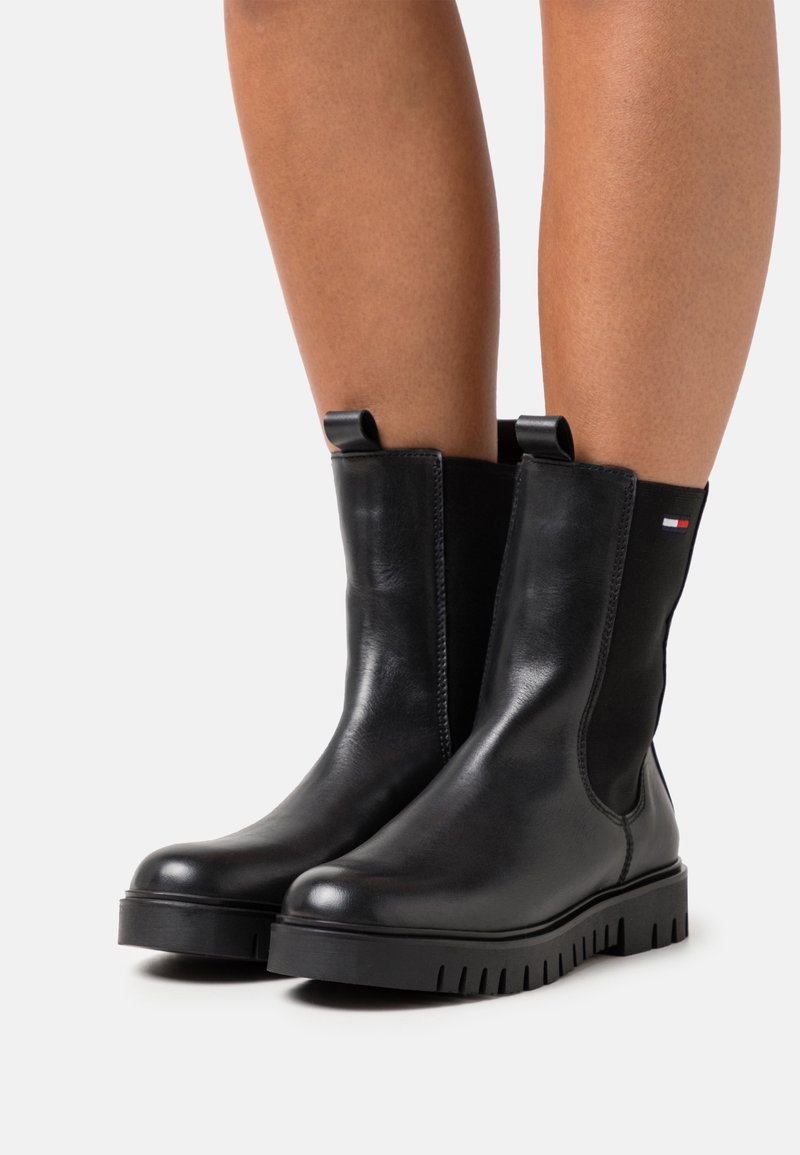 Bottes noires en cuir jusqu'au milieu du mollet avec languettes de traction, panneaux latéraux élastiques, semelles épaisses à crampons, portées sur des jambes nues sur fond blanc.