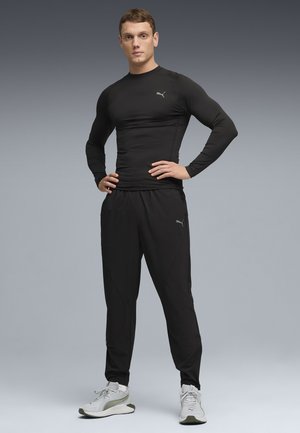 Uomo atletico con indosso una maglia nera a maniche lunghe, pantaloni neri e scarpe da ginnastica bianche, in piedi con le mani sui fianchi davanti a uno sfondo sfumato grigio.