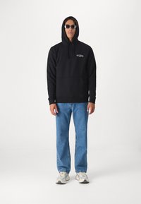 Black hoodie featuring a front kangaroo pocket and a logo design. Paired with light blue straight-leg jeans and beige sneakers.