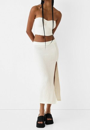 Kvinde iført en hvid, tekstureret, stropløs crop top og matchende højtaljet nederdel med et lårskørt, kombineret med sorte platåsandaler.