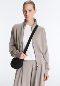 Žena oblečená v taupe bundě na zip a kalhotách s bílým tričkem, držící černou kabelku přes rameno na bílém pozadí.