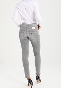 Jean ajustado de mezclilla gris con una cintura alta, bolsillos traseros y una etiqueta de cuero sintético. Combinado con una camisa blanca de botones y tacones negros.