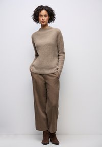 Beige neulepaita korkealla kauluksella, yhdistettynä leveisiin ruskeisiin housuihin ja ruskeisiin nilkkureihin. Tekstuuri on pehmeä ja pörröinen.