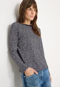 Granatowa bluzka z długim rękawem z białym geometrycznym wzorem. Luźny krój z okrągłym dekoltem, modelka ma rękę w kieszeni niebieskich jeansów.