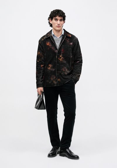 Jachetă de corduroy neagră cu modele abstracte colorate, asortată cu un pulover gri deschis, pantaloni negri și pantofi negri. Ține o geantă neagră.