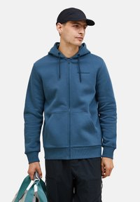 Svijetloplavi zip-up dukserica s kengurubažnom džepom, kapuljačom na šnuru i rebrastim manžetama. Logo otisnut na lijevoj strani prsnog koša. Tkanina izgleda glatko.