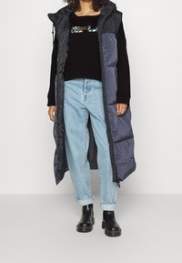 Dlouhá zateplená vesta v tmavě šedé barvě s kapucí, nošená přes černý svetr se flitrovým detailem. V kombinaci s světle modrými džínami a černými botami.