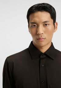 Bruine button-up shirt met een pointed collar, lange mouwen en zwarte knopen, gemaakt van een gladde, lichtgewicht stof. Getextureerde stiksels zichtbaar.