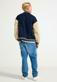 Marineblauwe en beige varsityjack met geribbelde kraag en manchetten, voorzien van een relaxte pasvorm, gecombineerd met lichtblauwe jeans en witte sneakers.