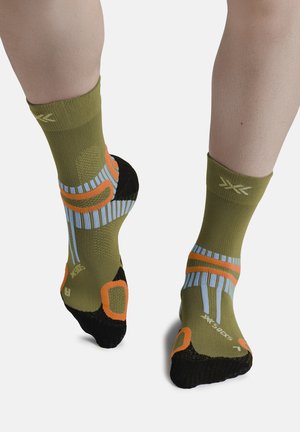 Voeten met groene sportsokken met zwarte hielen en tenen, met oranje en blauwe geometrische patronen, staand op een witte achtergrond.