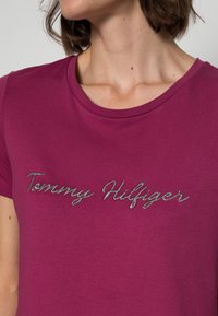 Fioletowa bawełniana koszulka z okrągłym dekoltem i krótkimi rękawami, ozdobiona srebrnym haftowanym napisem "Tommy Hilfiger".