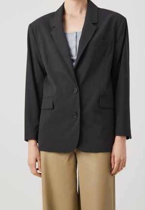 Blazer noir à deux boutons porté sur un haut bleu clair boutonné et un pantalon beige à jambes larges, sur une personne devant un fond uni.
