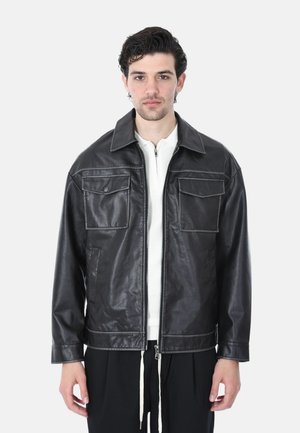 Giovane uomo con giacca di pelle nera con tasche a patta sul petto indossata sopra un maglione bianco con cerniera, in piedi davanti a uno sfondo bianco semplice.