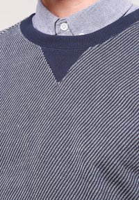 Navy knit sweater with a textured pattern, featuring a V-neck design and a light blue collared shirt underneath.