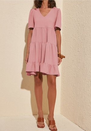Mujer que lleva un vestido rosa escalonado de manga corta con escote en V, pulseras marrones y sandalias marrones de tacón abierto, de pie en el interior.