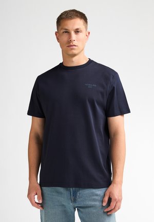 Giovane uomo con capelli corti che indossa una t-shirt blu navy con piccolo testo sul petto e jeans azzurri su uno sfondo semplice.