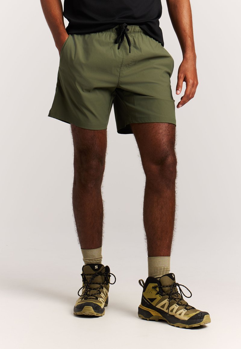 Olijfgroene korte broek met elastische tailleband en aantrekkoord. Draagt bijbehorende tankleurige wandelschoenen en beige sokken. Lichtgewicht, gladde stof.