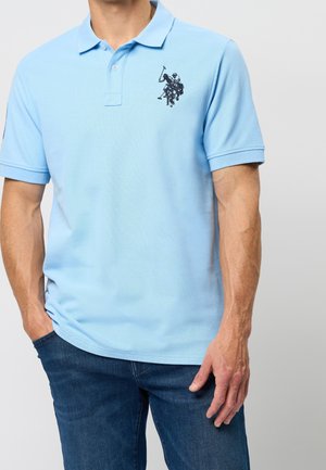 Mężczyzna w jasnoniebieskiej koszulce polo z czarnym, haftowanym logo zawodnika polo i niebieskich dżinsach, ręce w kieszeniach, na jednolitym tle.