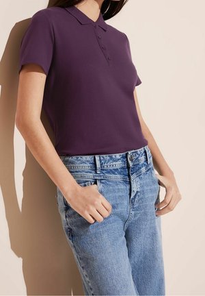 Femme portant un polo violet à manches courtes rentré dans un jean denim bleu clair taille haute, les mains près des poches.
