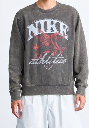 Osoba nosząca wyblakłą ciemnoszarą bluzę Nike z czerwonym graficznym skrzydlatym koniem i napisem „Nike athletics MCMLXXII”, w połączeniu z białymi szortami.