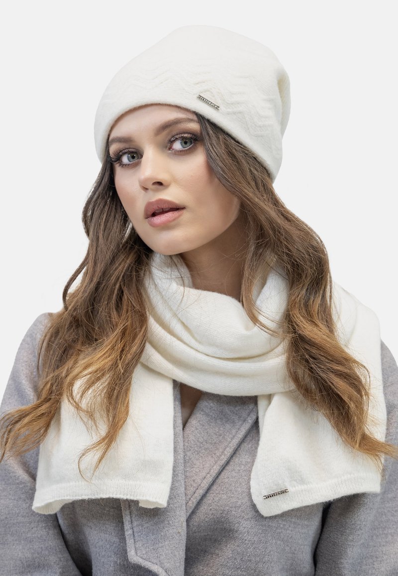 Witte gebreide beanie met zigzagpatroon en zilveren logo, gecombineerd met een bijpassende witte sjaal. Gladde textuur, zacht materiaal en een comfortabele uitstraling.