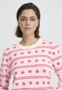 Pull rose avec un motif de cœur et des rayures horizontales. Fabriqué en tissu doux avec un col rond. Texture douce et design léger.