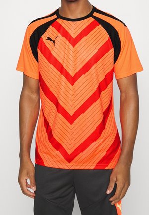 Uomo che indossa una maglietta atletica a maniche corte con un motivo a zigzag arancione brillante e rosso, con accenti neri, abbinata a pantaloni corti neri.