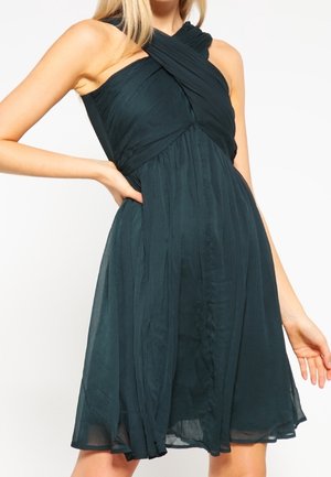 Czarna sukienka bez rękawów z krzyżującym się wycięciem typu halter i plisowaną spódnicą w kształcie litery A, noszona przez osobę z ręką na biodrze.