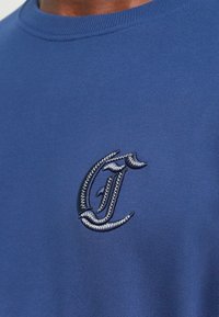 Tejido azul con una letra "C" estilizada bordada en hilo blanco y azul oscuro en la zona del pecho de una prenda.