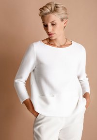White long-sleeve top with a round neck, textured fabric, and front pockets. Paired with light-colored trousers against a neutral background.