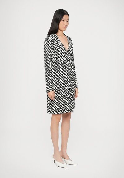 Μαύρο και λευκό φόρεμα με μοτίβο, μακριά μανίκια, V-λαιμόκοψη και δεμένη μέση. Λείο ύφασμα, μήκος μέχρι τη μέση του μηρού, συνδυασμένο με μυτερά λευκά τακούνια.