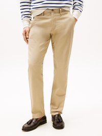 Beige Chinos mit glatter Textur, Frontverschluss, Seitentaschen und schmaler Passform, kombiniert mit dunkelbraunen Slippern.