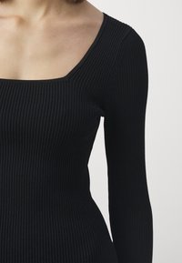 Top aderente a maniche lunghe nero con scollatura quadrata. Il tessuto presenta un aspetto texturizzato con linee verticali e una silhouette aderente.